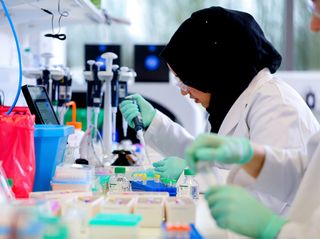 Scientist in a lab coat and hijab using a pipette in a laboratory, surrounded by scientific equipment and containers.